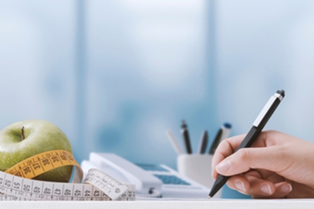 Professional nutritionist sitting at desk and writing a meal plan, measuring tape and apple